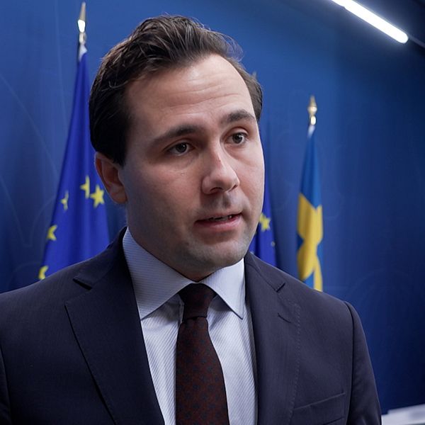 Benjamin Dousa (M) står framför en blå vägg prydd med svenska och europeiska flaggor. I bakgrunden syns en digital skärm.