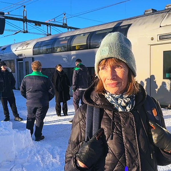 En person står på en snöig tågstation, medan ett tåg är placerat i bakgrunden och flera personer är synliga i närheten. Solen skiner och det finns en digital informationsskylt ovanför.