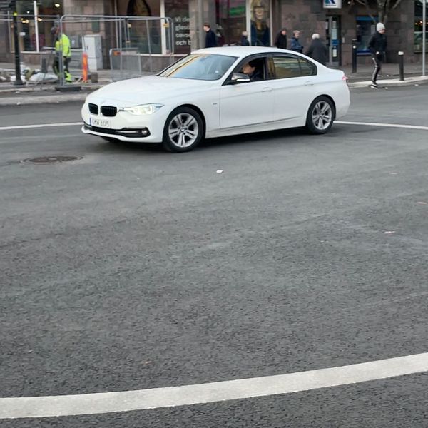 En vit bil svänger i en cirkulerande korsning, med en shoppinggata och byggnader i bakgrunden. Flera personer arbetar i närheten, och en annan bil är synlig längre fram i korsningen.