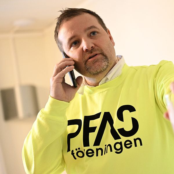 En person iförd en lysande gul tröja med texten ”PFAS-föreningen” står i en inomhusmiljö. Personen pratar i telefon och håller en glasbehållare i ena handen. Bakgrunden är neutral och svagt oskarp, vilket framhäver personen och deras handlingar.