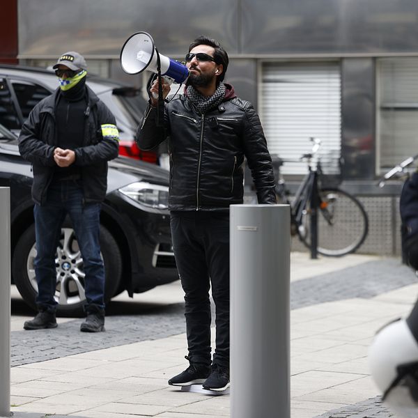 Salwan momika talar utomhus, polisen syns i förgrunden.