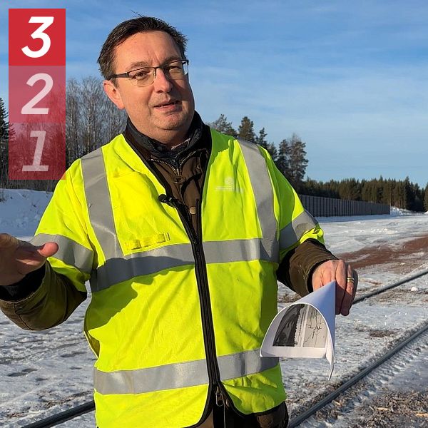 Person i gul reflexväst står vid en snöig väg med papper i handen, omgiven av skog.