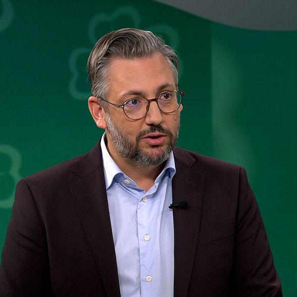 En manlig talare iklädd en mörk kostymjacka och en ljus skjorta står framför en grön bakgrund med blommönster. Bilden verkar vara tagen under en tv-sändning eller intervju. På skärmen syns texten ”Aktuellt.”
