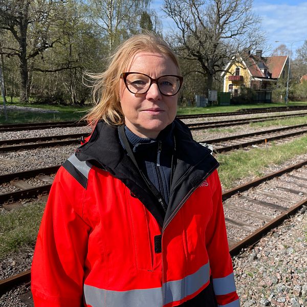 Person i röd reflexjacka står vid ett tågspår med hus och träd i bakgrunden.