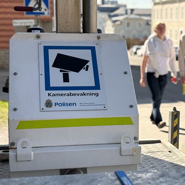 Skylt om kameraövervakning från polisen, tre personer promenerar i bakgrunden.