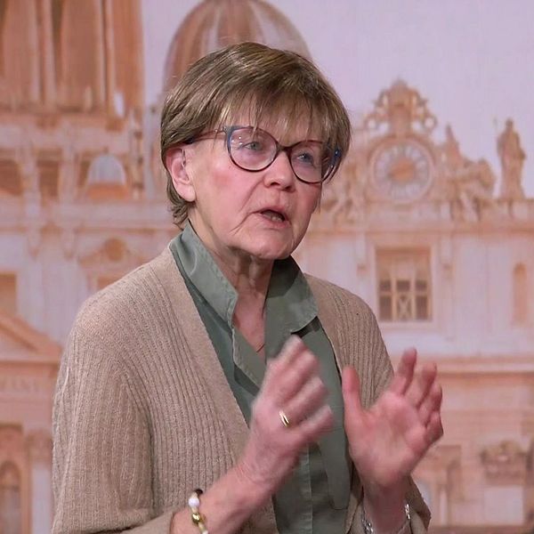 Person gestikulerar framför en bakgrund med klassisk arkitektur och kupolprydda byggnader. I bild syns Vatikankännaren Ulla Gudmundsson.
