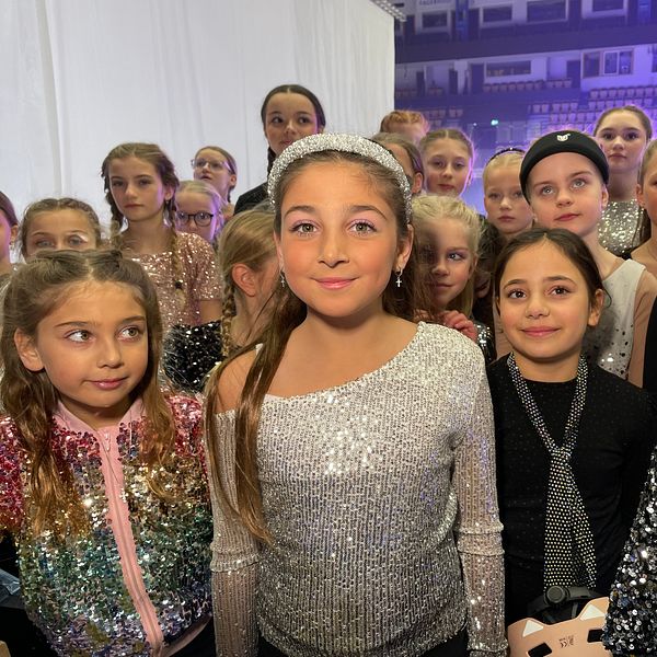 En grupp unga dansare klädda i glittriga kostymer samlade på en backstage-område inför en föreställning. De olika kostymerna varierar i färger och detaljer, vilket skapar en festlig atmosfär.