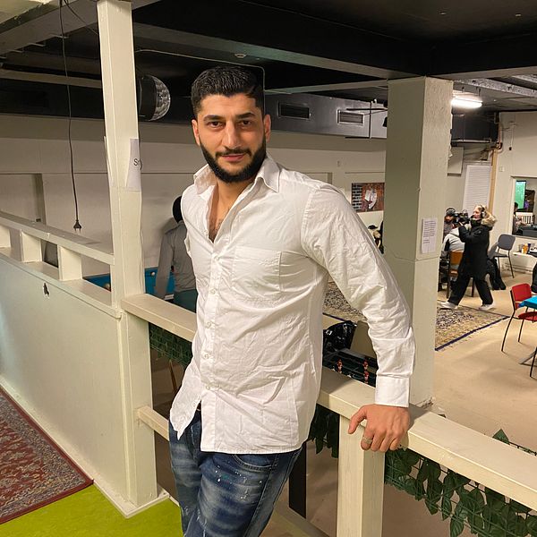 Nadjar Sheikho lutar sig mot en räcke i en lokal i Köping som används som fritidsgård. I bakgrunden syns människor som rör sig runt bord och stolar i olika färger.