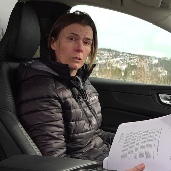 En kvinna i mörka kläder sitter i en bil. Hon tittar mot kameran och håller i en bunt papper