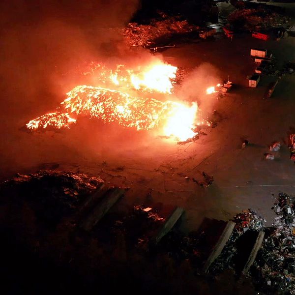 Stora lågor vid en brand på en återvinningscentral nattetid. Räddningstjänst på plats.