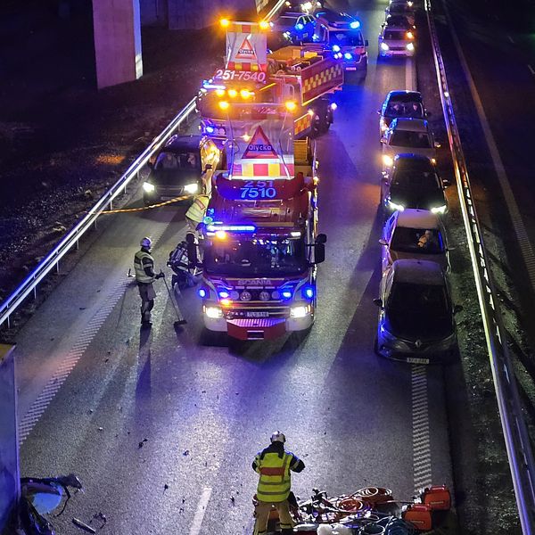Nattbild av en motorvägsolycka med flera räddningsfordon på plats. Brandmän i reflekterande kläder arbetar vid olycksplatsen, där flera bilar står uppradade längs vägen. Bärgning och säkerhetsåtgärder pågår.