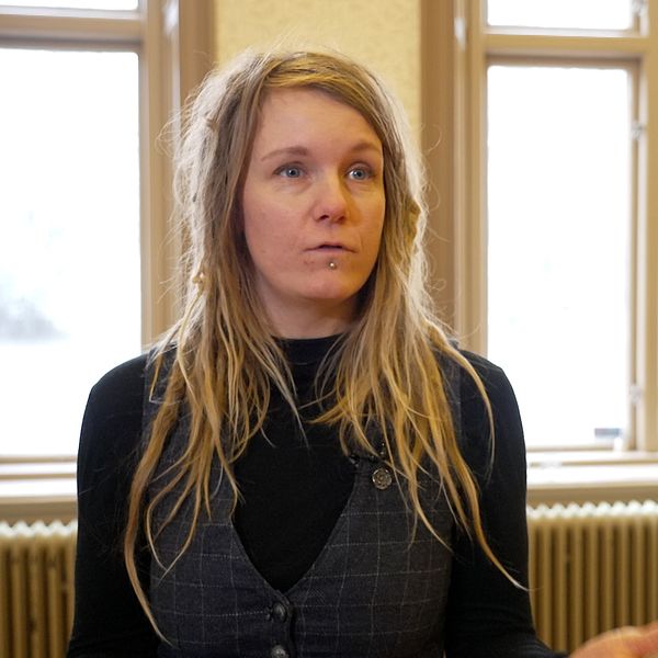 En person med långt blont hår står inomhus framför flera stora fönster och pratar.  Detaljerade inredningselement som en vägg med radiator och möbler är synliga i bakgrunden.