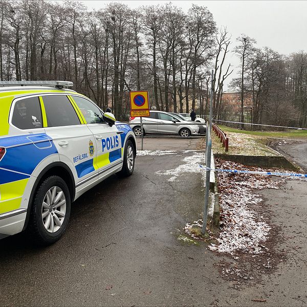 En polisbil står parkerad vid en avspärrad plats. Snö täcker delar av marken, och i bakgrunden syns en byggarbetsplats med ett blått skynke och en byggnadsställning.