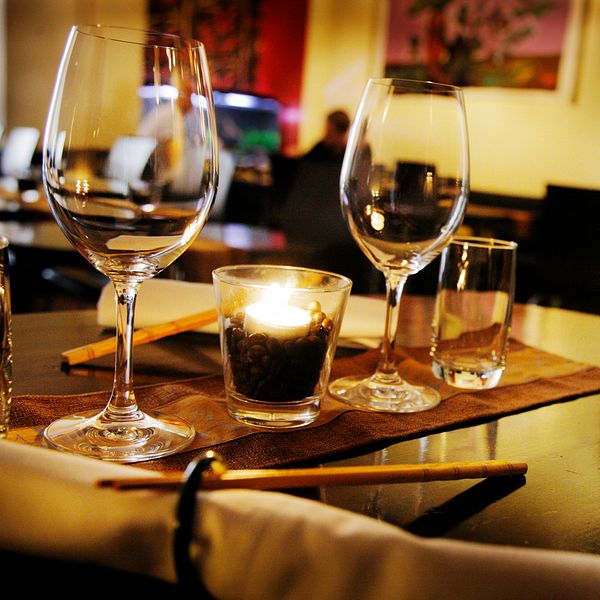 Ett dukat bord i en restaurang med tre vinglas, ett ljus i en glasbehållare med mörka stenar, och en liten shotglas. I bakgrunden syns oskarpa silhuetter av gäster och en bar.