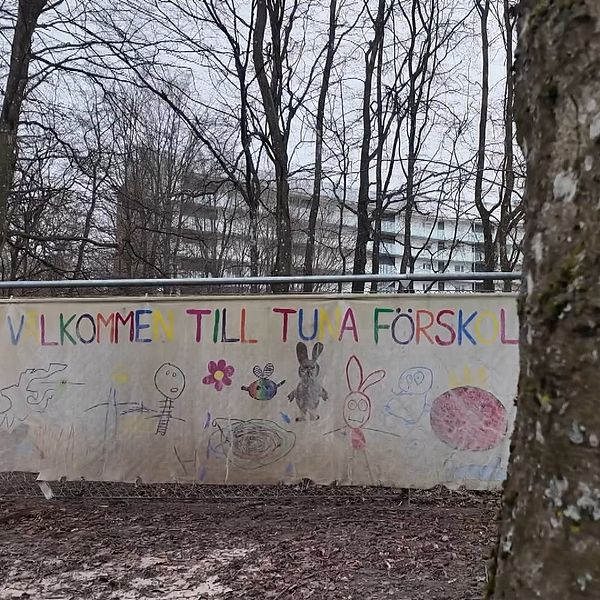 Träd i förgrunden, lakan med text ”VÄLKOMMEN TILL TUNA FÖRSKOLA” i bakgrunden.