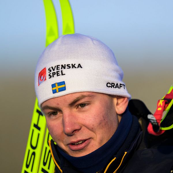 En person står med skidor och stavar, iförd en ljus mössa med texten ”SVENSKA” samt en jacka. Skidorna och stavarna hålls i ena handen, och bakgrunden är suddig men visar en kall och klar miljö.