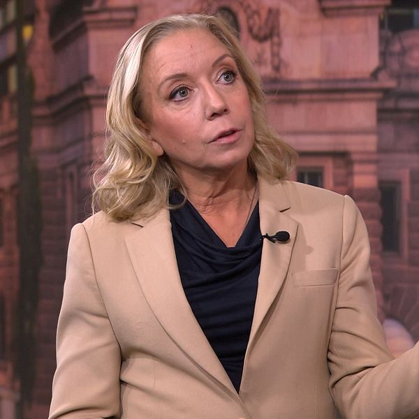 Person i beige kavaj gestikulerar framför byggnad.