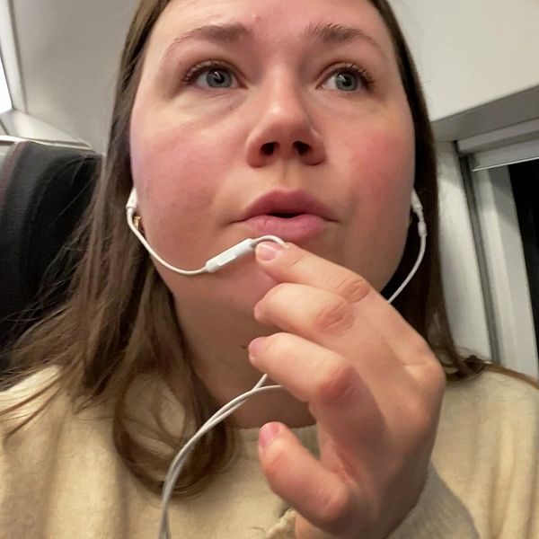 Kvinna i brunt hår sitter i en tågkupé, i tågets tysta avdelning, och håller en hörlur i handen. Bakgrunden visar fönstret i tågkupén, med svagt ljus och en del av järnvägsstationen synlig utanför. Personen har på sig en ljus tröja och ser ut att vara upptagen med att lyssna på något.
