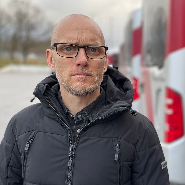 En person iförd en svart jacka står framför flera röda och vita bussar i en parkeringsplats. Det är en molnig dag med en grönskande bakgrund. Bussarna är i fokus och skapar en känsla av en transport- eller reseverksamhet.