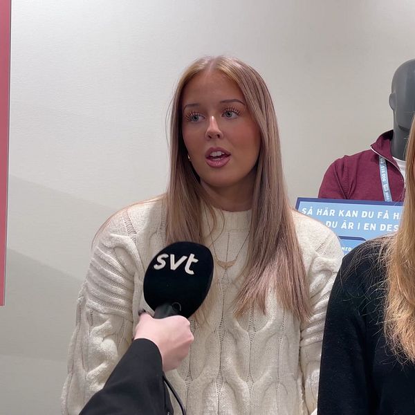 Två personer står framför en vägg med information. . Väggen har olika meddelanden och symboler relaterade till ett ämne, troligen kopplat till utbildning eller information.