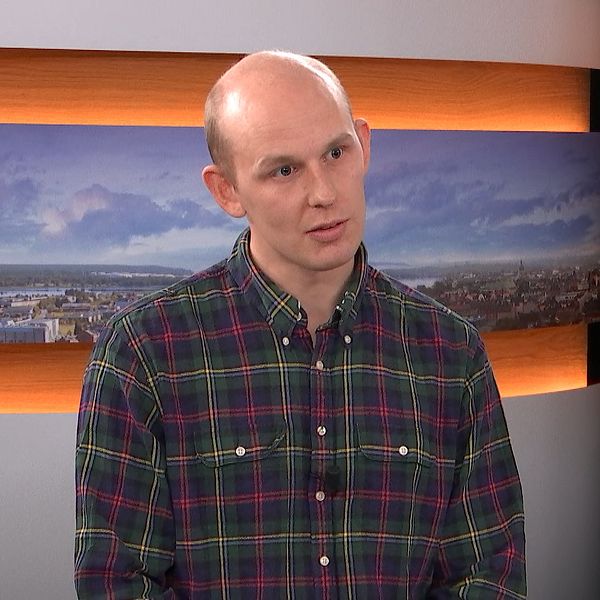 En man står i en intervju i en studio, klädd i en rutig skjorta. I bakgrunden syns panoramabilder av en stad med grönska och vatten. Belysningen är mjuk och ger en professionell atmosfär.