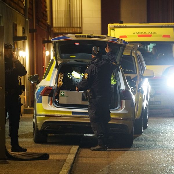 Poliser står vid en öppen baklucka på en polisbil i mörker, med en ambulans synlig i bakgrunden.