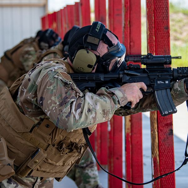 En soldat i kamouflageuniform och skyddsutrustning tar sikte med ett automatvapen vid en träningsanläggning. Bakom honom står fler soldater, och bakgrunden visar en mån av röd färg och grön vegetation.