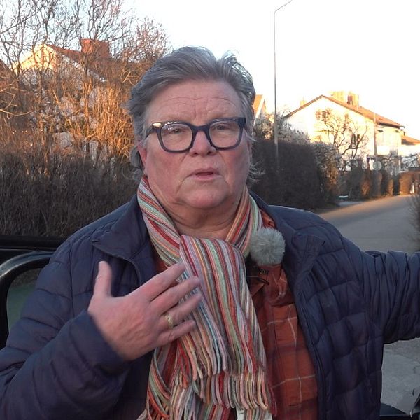 En person står brevid en bil och gestikulerar med handen, iklädd en blå jacka och en randig halsduk, mot en bakgrund av bostadshus och en vägskylt.