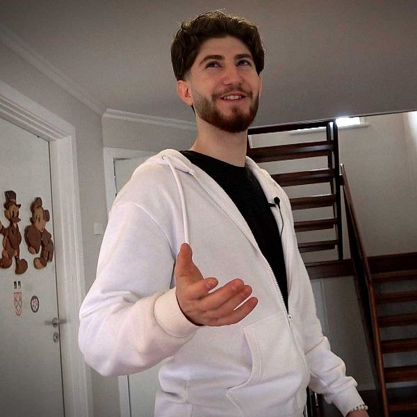 Alexander Bisso i vit hoodie står i hallen framför trappa, räcker ut handen mot kameran.