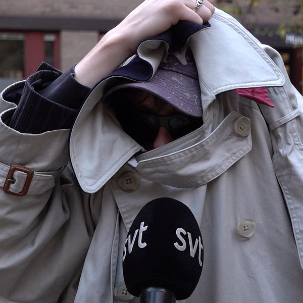 En person i en beige trench coat håller upp kappan framför sig medan de talar. En mikrofon med logotypen för SVT syns i nederkanten av bilden. Bakgrunden visar en del av en byggnad med röda fönster och omgivande natur med höstlöv.