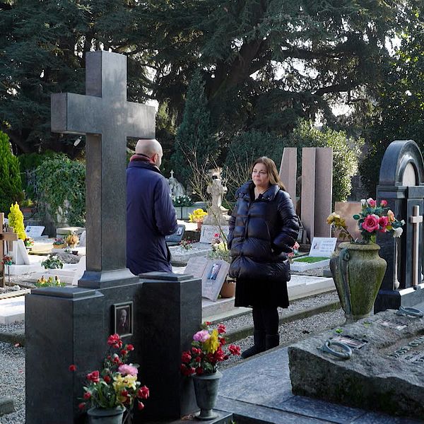 Två personer står vid gravstenar på en kyrkogård omgivna av blommor och grönska.