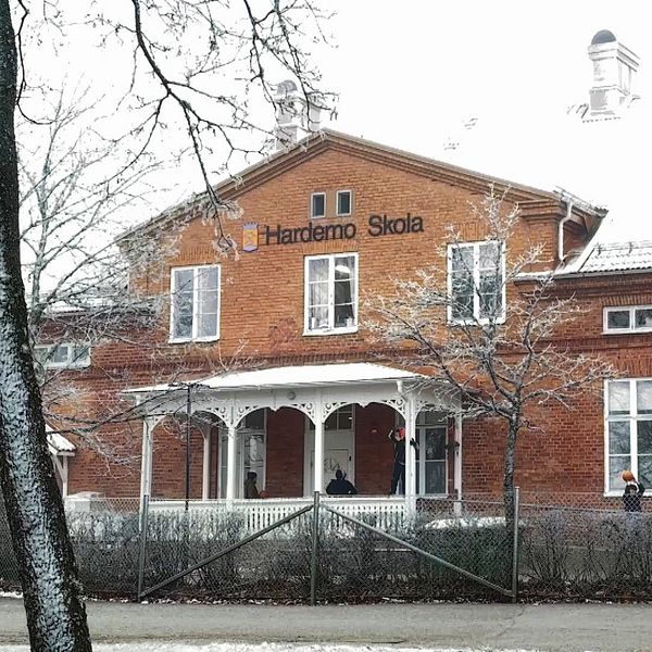 Röd tegelbyggnad som utgör Hardemo Skola, omgiven av snötäckta träd och en vinterlandskap. Byggnaden har en traditionell stil med en veranda och flera fönster. En del av skolan är annoterad med skolans namn.
