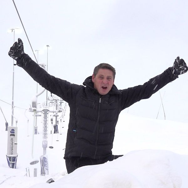 En glad man sitter i en snöig skidbacke framför en skidlift