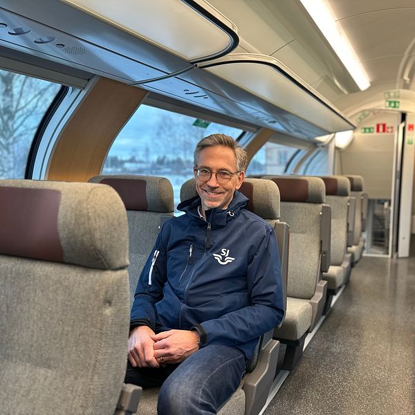 En person sitter på en tom, modern tågkupé med grå tygklädda säten. Tågkupén är ljust upplyst.