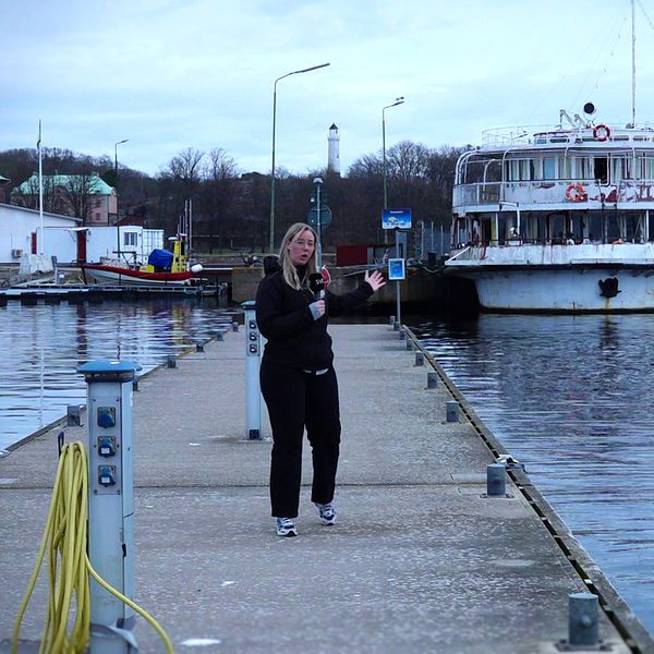 Reporter Cajsa Bengtsson går på brygga i Karlskrona, med Studentbåten förtöjd i bakgrunden. Mulet väder.