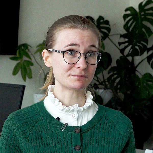 Forskaren Emma Samuelsson är klädd i en grön tröja med vit krage står framför en grön växt och en dator i en kontorsmiljö. Bakgrunden inkluderar svarta kontorsstolar och en stor växt som ger en hemtrevlig känsla i rummet.