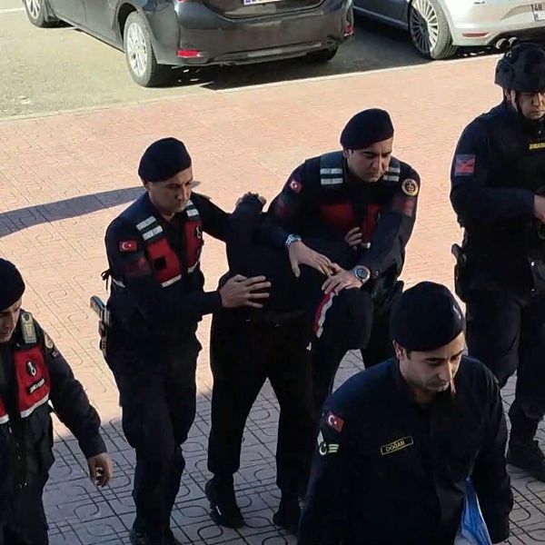 Flera poliser i uniform eskorterar en person i handfängsel. En av poliserna håller ett automatvapen. Scenen utspelar sig utomhus på en trottoar vid parkerade bilar.
