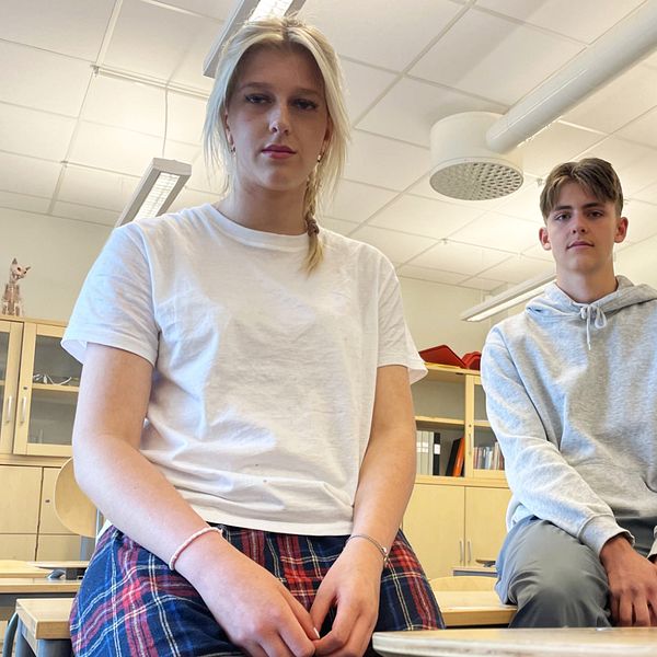 En blond kille och tjej sitter bredvid varandra på en skolbänk i ett klassrum och kollar in i kameran.