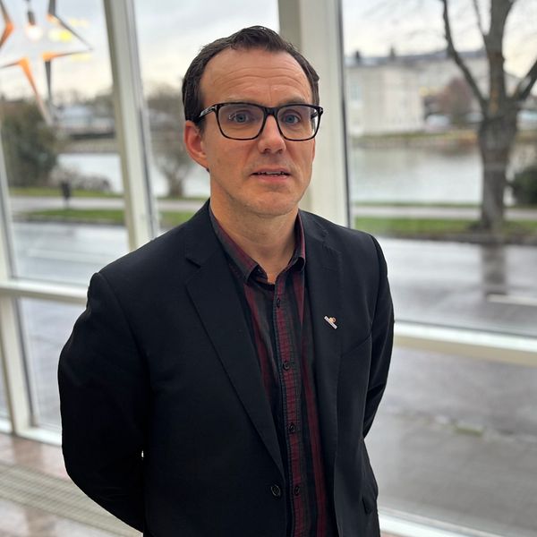 En person står framför ett stort fönster med utsikt över en flod och en stadsmiljö. Det dämpade ljuset inomhus kontrasterar med den ljusare omgivningen utanför. Det är en modern inredning med stiliga detaljer.
