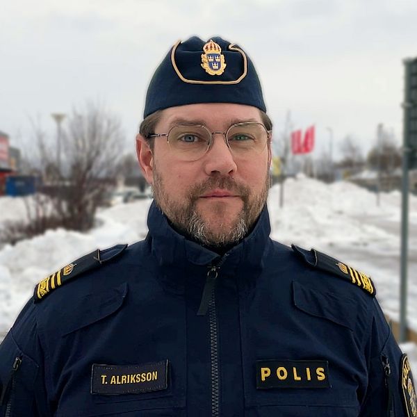 En polis i uniform står i ett snöigt stadsområde med en väg och flaggor i bakgrunden.