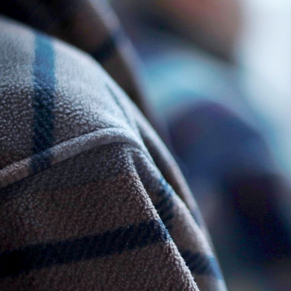 Närbild av en persons axel iklädd en rutig fleecejacka med fokus på tygets textur medan bakgrunden är oskarp och visar vaga konturer av ytterligare personer eller föremål.
