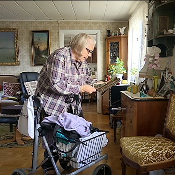Äldre kvinna med rullator läser papper i ett hemtrevligt vardagsrum.