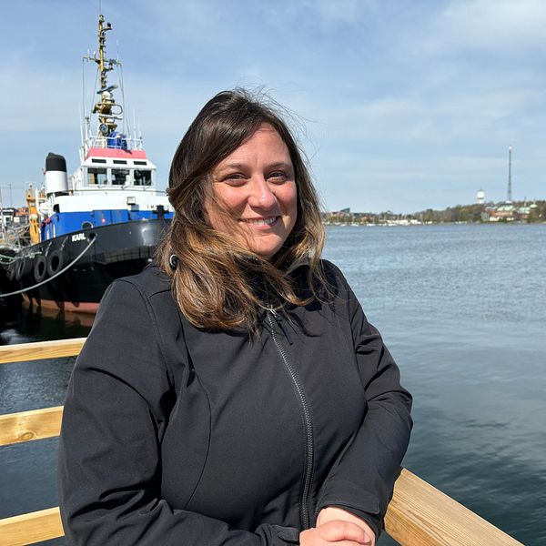 Elena Peverada står i svart jacka står vid vattnet, fartyg och kuststad syns i bakgrunden.