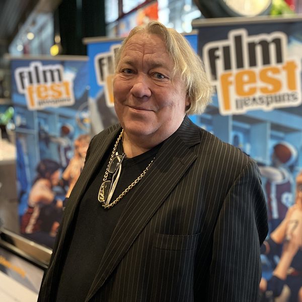 Leif Andrée iklädd en svart randig kavaj står framför en bakgrund med affischer för ”Filmfest” som visar olika scener från filmfestivalen. Affischerna har färgglada bilder av filmiska element och texten ”film fest”. Miljön ser ut att vara ett evenemangsutrymme.