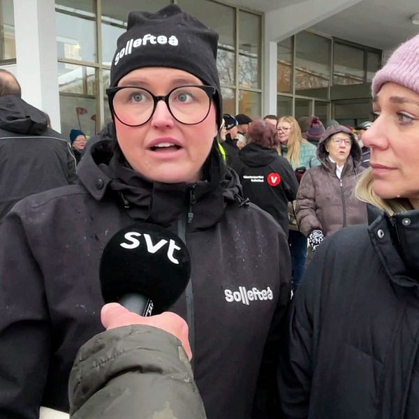 Två personer står i en grupp utanför en byggnad, där en av dem håller en mikrofon med SVT-logotypen. I bakgrunden syns en samling människor och en del vinterklädda personer.