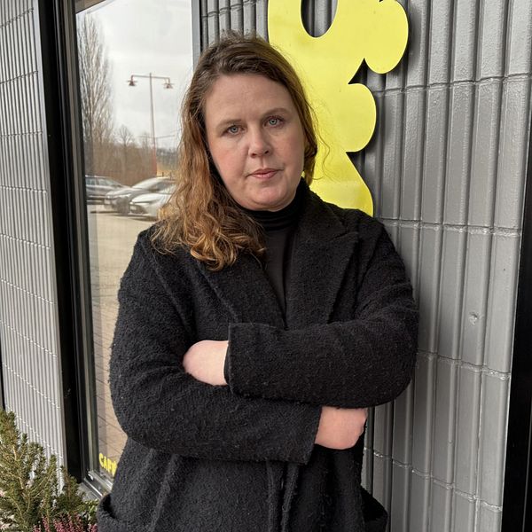 En person med axellångt hår står framför en butiksentré med glasdörrar, klädd i en svart jacka. I bakgrunden syns en del av en gul skylt och öppettiderna för butiken.