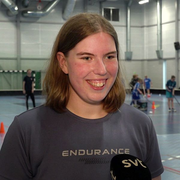 Person intervjuas i idrottshall där människor spelar innebandy i bakgrunden. SVT-mikrofon synlig.