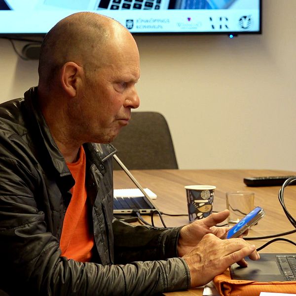 medelålders man med rakat huvud sitter med mobiltelefon framför sin dator i ett konferensrum
