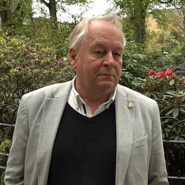 Sven Andersson i grå kavaj står vid ett svart staket i grönskande parkmiljö.