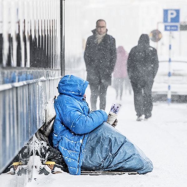 EU-migrant sitter och tigger, har vinterkläder på sig och det snöar.